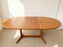 Load image into Gallery viewer, Image 10 of 28: An oval wooden dining table with a warm medium-brown finish, featuring a central seam and a four-leg base, set against a white brick wall and light-colored carpet.
