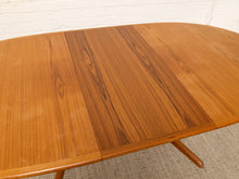 Load image into Gallery viewer, Image 23 of 28: A close-up of a wooden dining table top with three visible wood-grain panels and part of a curved armrest, photographed indoors against a white brick wall and light carpet.
