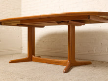 Load image into Gallery viewer, Image 25 of 28: A mid-century modern wooden dining table with a warm medium-brown finish, featuring a drop-leaf design and a wide pedestal base, set against a white brick wall and light carpet.
