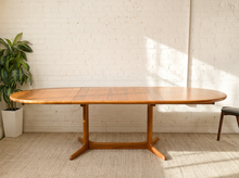 Load image into Gallery viewer, Image 11 of 28: A mid-century modern wooden dining table with an oval top and three-star base, set against a white brick wall with a potted plant on the left.
