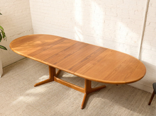 Load image into Gallery viewer, Image 12 of 28: An oval wooden dining table with a warm medium-brown finish, featuring a central stretcher base and extendable leaf-like side panels, set against a white brick wall and beige carpet.
