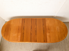Load image into Gallery viewer, Image 13 of 28: An oval wooden table with a warm honey-toned wood grain finish, photographed from above against a white brick wall and light carpet.
