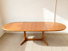 Load image into Gallery viewer, Image 14 of 28: An oval wooden dining table with a warm medium-brown finish and a star base, set against a white brick wall and light-colored rug.
