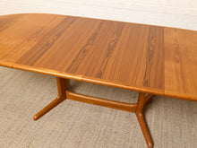 Load image into Gallery viewer, Image 26 of 28: A mid-century modern wooden dining table with a warm honey-toned teak wood grain, featuring a curved apron base and extendable leaf panels, set on a textured beige rug against a white brick wall.
