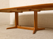 Load image into Gallery viewer, Image 28 of 28: A mid-century modern wooden dining table with a warm honey-toned finish, featuring a slatted top and a wide T-shaped pedestal base, photographed in a room with a white brick wall and textured carpet.
