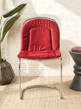 Load image into Gallery viewer, Image 4 of 15: A mid-century modern accent chair with a red velvet cushioned seat and backrest, supported by a curved chrome metal frame, set against a white brick wall.
