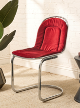 Load image into Gallery viewer, Image 5 of 15: A modern side chair with a plush red velvet seat and back cushion, supported by a curved polished chrome metal frame, set against a white brick wall background.
