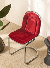 Load image into Gallery viewer, Image 6 of 15: A modern cantilever chair with a plush red velvet cushioned seat and backrest, supported by a curved polished chrome metal frame, set on a beige woven rug against a white brick wall.
