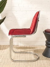 Load image into Gallery viewer, Image 7 of 15: A modern cantilever chair with a red upholstered seat and backrest, supported by a curved chrome metal base, set against a white brick wall background.
