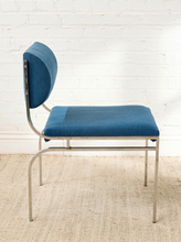 Load image into Gallery viewer, Image 5 of 14: A mid-century modern dining chair with blue upholstered seat and backrest, supported by a chrome tubular steel frame, set against a white brick wall and a beige woven rug.
