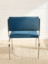Load image into Gallery viewer, Image 6 of 14: A mid-century modern dining chair with blue upholstered seat and backrest, supported by a polished chrome metal frame, set against a white brick wall and a beige woven rug.
