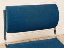 Load image into Gallery viewer, Image 7 of 14: A close-up of a mid-century modern chair with a curved backrest and seat upholstered in textured blue fabric, supported by a chrome tubular steel frame against a white brick wall.

