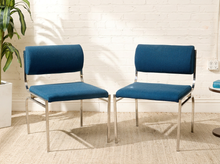 Load image into Gallery viewer, Image 14 of 14: Two modern dining chairs with blue upholstered seats and backs and chrome metal frames, set against a white brick wall with a potted plant on the left.
