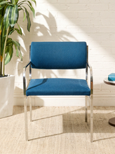 Load image into Gallery viewer, Image 2 of 15: Vintage dining armchair featuring a navy blue upholstered seat, curved cylindrical padded backrest, open-back design, polished chrome armrests, and slender angled metal legs.
