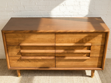 Load image into Gallery viewer, Image 6 of 16: Mid-century modern wooden chest of drawers with six drawers, featuring integrated horizontal pull handles and tapered legs, set against a white brick wall.
