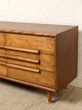 Load image into Gallery viewer, Image 7 of 16: Mid-century modern wooden dresser with curved drawer pulls and tapered legs, shown against a white brick wall and textured rug.
