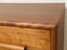 Load image into Gallery viewer, Image 11 of 16: Close-up of a wooden dresser or chest with a warm medium-brown finish, showing the top surface, drawer front, rounded corner trim, and part of a curved drawer pull against a light wall.
