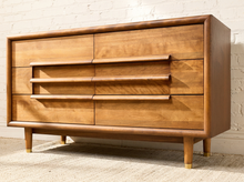 Load image into Gallery viewer, Image 12 of 16: Mid-century modern wooden dresser with six drawers, curved horizontal pull handles, and tapered legs, shown against a white brick wall and light rug.
