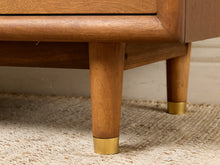 Load image into Gallery viewer, Image 13 of 16: Close-up of a mid-century modern wooden furniture piece, showing the warm brown wood grain of the legs and drawer front, accented with brass metal caps on the leg ends.

