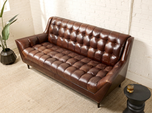 Load image into Gallery viewer, Image 3 of 13: A mid-century modern brown leather tufted loveseat sofa in a styled room setting with a white brick wall, beige carpet, a potted plant, and a small side table.
