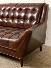 Load image into Gallery viewer, Image 5 of 13: A close-up of a dark brown tufted leather sofa with angled arms and dark tapered legs, set against a white brick wall and textured beige rug.
