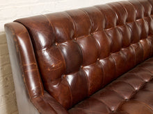 Load image into Gallery viewer, Image 7 of 13: Close-up of a brown tufted leather sofa with rolled arms and a distressed finish, set against a white brick wall.
