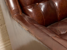 Load image into Gallery viewer, Image 8 of 13: Close-up of a brown leather armchair showing the armrest, tufted back cushion with a button, and worn edge of the leather arm against a light neutral wall.
