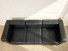 Load image into Gallery viewer, Image 4 of 14: A three-seat black leather sofa with a low-profile rectangular design, shown from a top-down angle against a white brick wall and light-colored woven rug.
