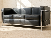 Load image into Gallery viewer, Image 13 of 14: A mid-century modern black leather sofa with a polished chrome steel frame, positioned in front of a white brick wall with natural light casting shadows.

