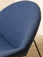 Load image into Gallery viewer, Image 8 of 17: Close-up of a blue upholstered chair seat with a textured fabric, showing the curved backrest and a portion of the black metal leg against a light brick wall and beige floor.
