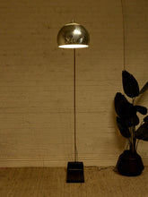 Load image into Gallery viewer, Image 6 of 23: A modern floor lamp with a polished metallic dome shade and a slim black stand, illuminated against a warm-toned brick wall next to a potted plant.
