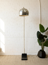Load image into Gallery viewer, Image 5 of 23: A modern floor lamp with a polished silver dome shade, a slim gold-toned stem, and a black rectangular base, standing on a woven rug against a white brick wall next to a potted green plant.
