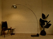 Load image into Gallery viewer, Image 8 of 23: A modern floor lamp with a long curved metallic arm and a reflective dome shade stands in a room with a brick wall, next to a black chair and a potted plant.
