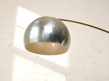 Load image into Gallery viewer, Image 9 of 23: A modern floor lamp with a large, dome-shaped polished chrome shade and a curved brass-colored metal arm, set against a white brick wall with soft window light casting shadows.
