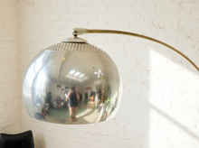 Load image into Gallery viewer, Image 11 of 23: A modern arched floor lamp with a large, highly polished silver dome shade and a curved brass-colored metal arm, set against a white brick wall.
