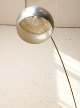 Load image into Gallery viewer, Image 13 of 23: A modern floor lamp with a large spherical shade in brushed silver and a long curved brass-colored metal stem, photographed against a white painted brick wall with sunlight casting shadows.
