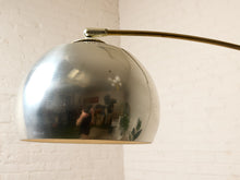 Load image into Gallery viewer, Image 15 of 23: A close-up of a modern floor lamp with a large, polished silver dome shade and a brass-colored curved arm, set against a white brick wall.
