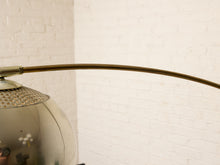 Load image into Gallery viewer, Image 17 of 23: A close-up of a modern floor lamp featuring a curved brass metal arm and a reflective, dome-shaped glass shade, set against a white brick wall background.

