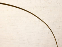 Load image into Gallery viewer, Image 18 of 23: A close-up of a curved metallic floor lamp arm in a warm gold/brass tone, set against a white-painted brick wall.
