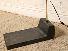 Load image into Gallery viewer, Image 20 of 23: A vintage-style floor lamp with a thick, rectangular black cast-iron base and a slim brass pole, resting on a textured beige carpet against a white brick wall.

