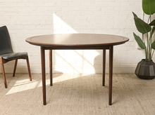 Load image into Gallery viewer, Image 2 of 12: Modern round dining table featuring a warm walnut-tone wood grain finish, subtly segmented circular tabletop border, softly rounded edges, and slender angled tapered legs.
