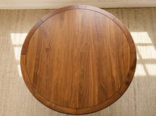 Load image into Gallery viewer, Image 4 of 12: Modern round dining table featuring a warm walnut-tone wood grain finish, subtly segmented circular tabletop border, softly rounded edges, and slender angled tapered legs.
