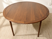 Load image into Gallery viewer, Image 5 of 12: A round wooden dining table with a warm medium-brown walnut finish, featuring four tapered legs and a visible leaf extension seam on the tabletop, set on a textured beige rug against a white brick wall.
