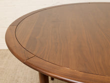Load image into Gallery viewer, Image 6 of 12: Close-up of a round wooden dining table with a warm medium-brown finish and visible wood grain, showing part of the tabletop and leg against a light neutral background.
