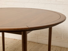 Load image into Gallery viewer, Image 8 of 12: A close-up of a round wooden dining table with a warm medium-brown finish, showing the tabletop seam and legs against a white brick wall and light rug.
