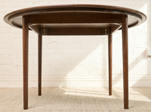 Load image into Gallery viewer, Image 9 of 12: A round wooden dining table with a dark brown finish and visible metal hardware on the underside, set against a white brick wall and light-colored floor.

