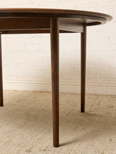Load image into Gallery viewer, Image 10 of 12: A close-up of a dark brown wooden dining table with tapered legs, set against a white brick wall and a light beige woven rug.
