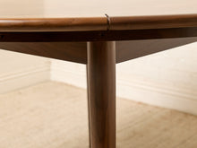 Load image into Gallery viewer, Image 11 of 12: Close-up of a wooden dining table showing the underside of the tabletop, a central support leg, and part of the room background with carpet and baseboard.
