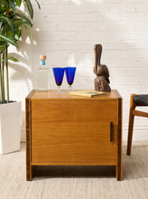 Load image into Gallery viewer, Image 1 of 17: Vintage mid-century modern rectangular wooden cabinet featuring a honey-brown wood grain finish, single flat-panel door, dark vertical metal pull, and contrasting darker wood trim along both sides.
