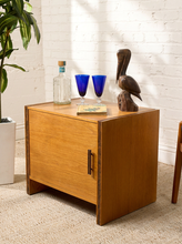 Load image into Gallery viewer, Image 2 of 17: Vintage mid-century modern rectangular wooden cabinet featuring a honey-brown wood grain finish, single flat-panel door, dark vertical metal pull, and contrasting darker wood trim along both sides.
